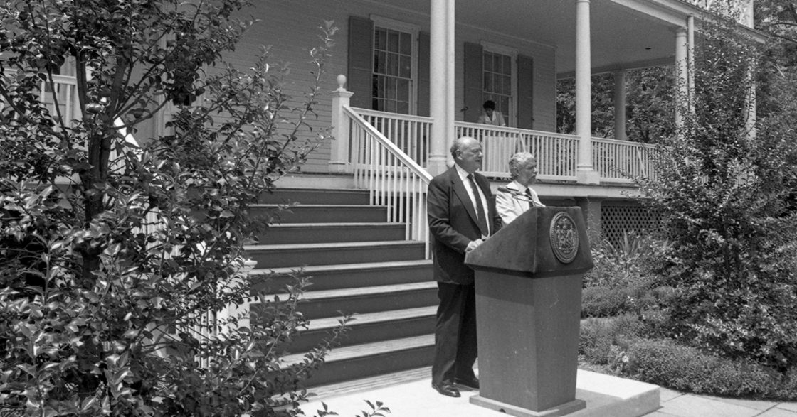 A Black-and-White photo of Mayor Edward I. Koch and Parks Commissioner Henry J. Stern stand at a wooden podium in front of the steps of Gracie Mansion.