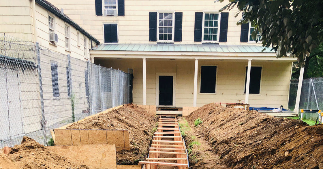 King Manor, a yellow house with black shutters, is pictured with piles of dirt in the yard. The exposed ground shows the infrastructure built for the HVAC reconstruction preservation project.