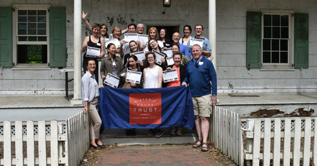 A group of museum volunteers poses for a photo on the steps of a historic house while holding a flag with the red-and-white Historic House Trust logo.