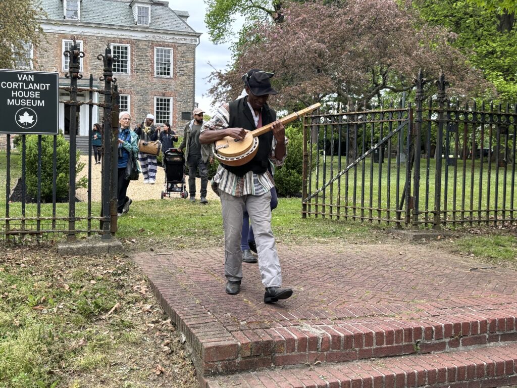 Man playing the banjo walking out of the Van Cortlandt House Museum grounds through the front gate.