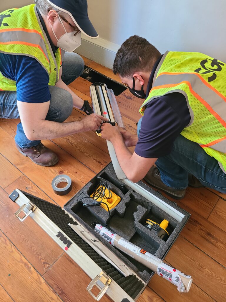 Two men in yellow reflective vest using complex equipment near a brown florr.