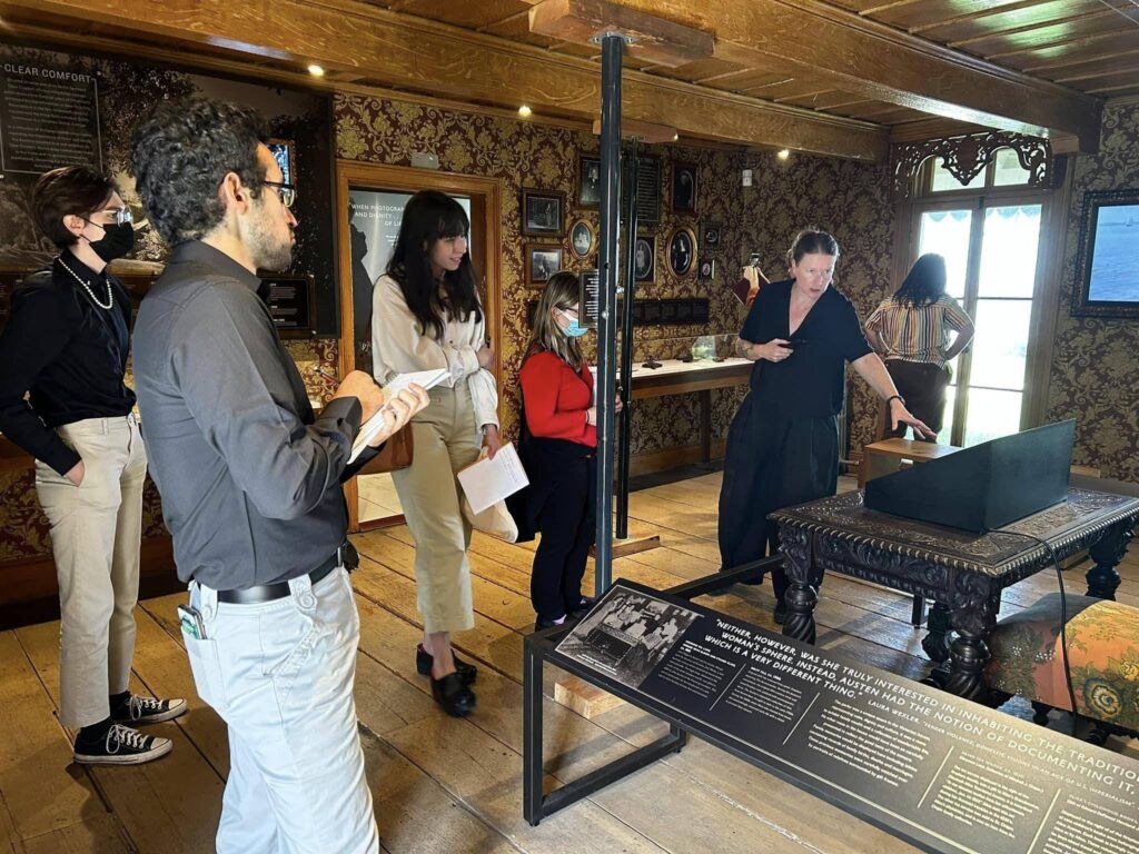 Woman in black pointing to display of objects in front of a group of students. 