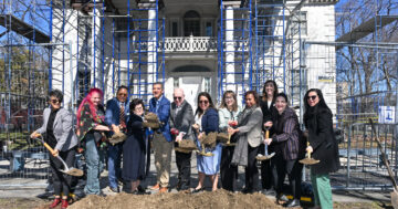 12 people throwing shovels of dirt outside of a white historic house with blue scaffolding