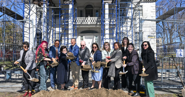 12 people throwing shovels of dirt outside of a white historic house with blue scaffolding