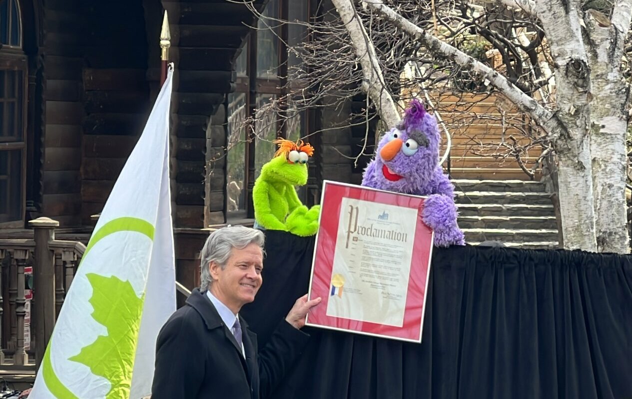 Man with white hair holding a framed document with one purple and one green puppet. Standing in front of a white and green flag, and a black curtain. 